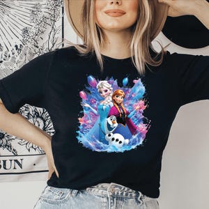 Puede incluir: Camiseta negra con un gr&aacute;fico colorido de Elsa, Anna y Olaf de la pel&iacute;cula Frozen. El dise&ntilde;o incluye salpicaduras azules, rosas y moradas al estilo acuarela. La camiseta tiene un cuello redondo cl&aacute;sico.