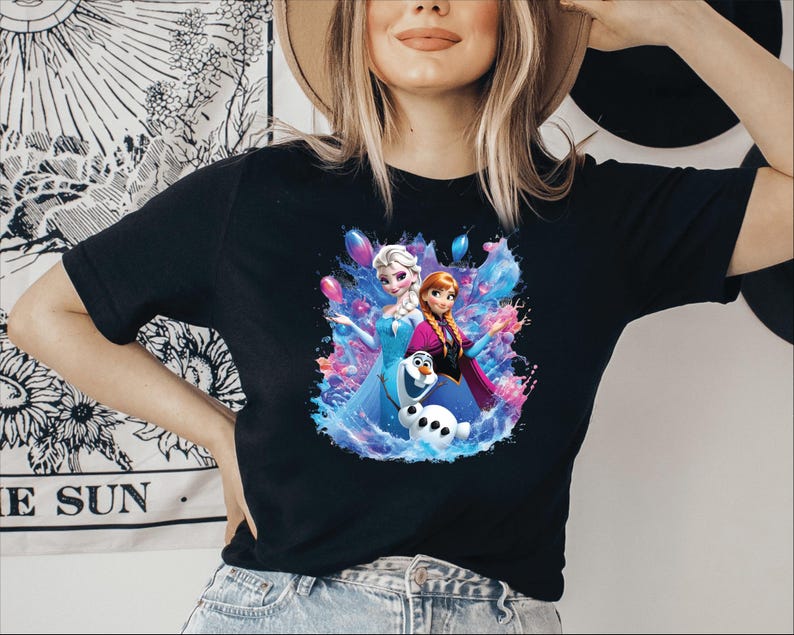 Puede incluir: Camiseta negra con un gr&aacute;fico colorido de Elsa, Anna y Olaf de la pel&iacute;cula Frozen. El dise&ntilde;o incluye salpicaduras azules, rosas y moradas al estilo acuarela. La camiseta tiene un cuello redondo cl&aacute;sico.