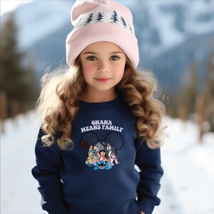 K&ouml;nnte beinhalten: Ein junges Kind tr&auml;gt einen marineblauen Pullover mit dem Text "OHANA MEANS FAMILY" und einer Grafik von Disney-Figuren. Die Person tr&auml;gt auch eine hellrosa M&uuml;tze mit einem wei&szlig;en Baummuster. Der Hintergrund ist eine verschneite Landschaft.