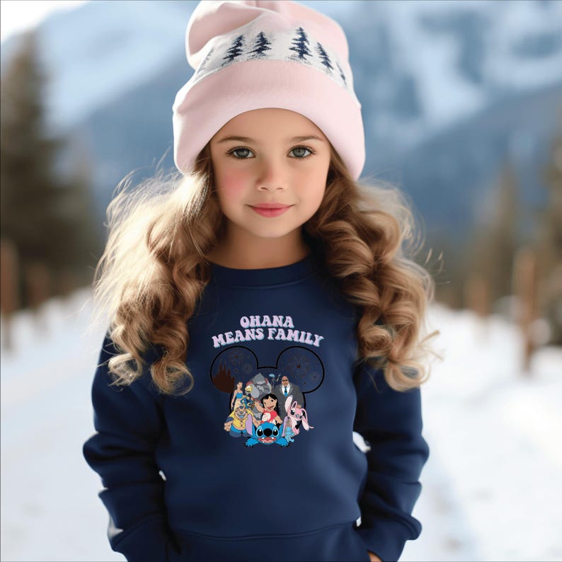 K&ouml;nnte beinhalten: Ein junges Kind tr&auml;gt einen marineblauen Pullover mit dem Text "OHANA MEANS FAMILY" und einer Grafik von Disney-Figuren. Die Person tr&auml;gt auch eine hellrosa M&uuml;tze mit einem wei&szlig;en Baummuster. Der Hintergrund ist eine verschneite Landschaft.