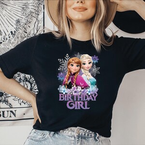 K&ouml;nnte beinhalten: Schwarzes T-Shirt mit einer Grafik von Anna und Elsa, animierten Prinzessinnen, umgeben von Schneeflocken und Blumen. Der Text "BIRTHDAY GIRL" steht darunter in Lila. Das T-Shirt wird von einer Person getragen.