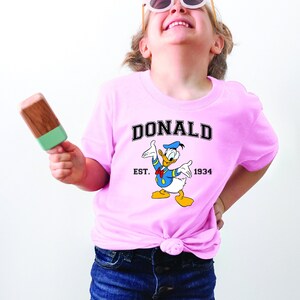 May include: A pink t-shirt with a cartoon image of Donald Duck. The shirt has the text "DONALD" in a black arch above the image and "EST. 1934" below the image.