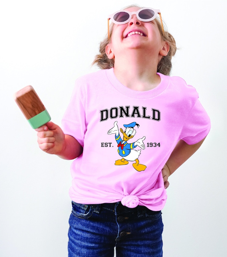 May include: A pink t-shirt with a cartoon image of Donald Duck. The shirt has the text "DONALD" in a black arch above the image and "EST. 1934" below the image.