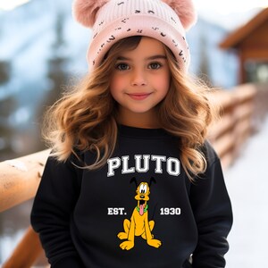 Puede incluir: Una sudadera negra con una imagen de dibujos animados de Pluto, el perro de Disney. La sudadera tiene el texto "PLUTO" en letras blancas sobre la imagen y "EST. 1930" en letras blancas debajo de la imagen.