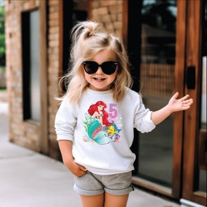 May include: A white long-sleeved shirt with a graphic of Ariel the mermaid from The Little Mermaid, Flounder, and the number 5. The shirt is worn by a child wearing black sunglasses.