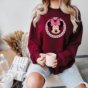 Puede incluir: Una sudadera color burdeos con un c&iacute;rculo a cuadros blancos que presenta una imagen de dibujos animados de Minnie Mouse con un lazo rosa.