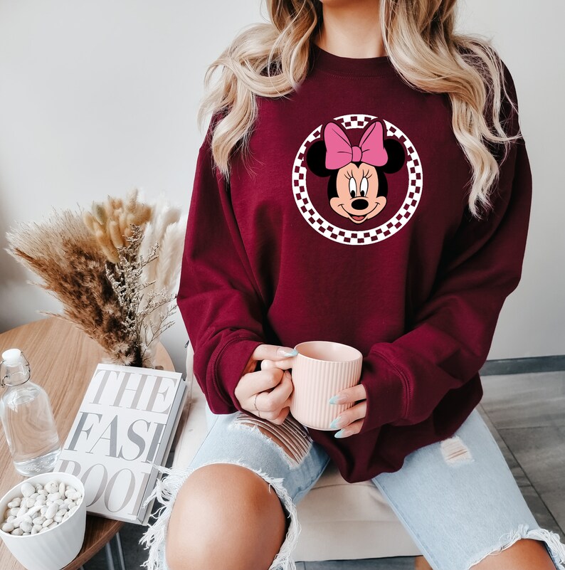 Puede incluir: Una sudadera color burdeos con un c&iacute;rculo a cuadros blancos que presenta una imagen de dibujos animados de Minnie Mouse con un lazo rosa.