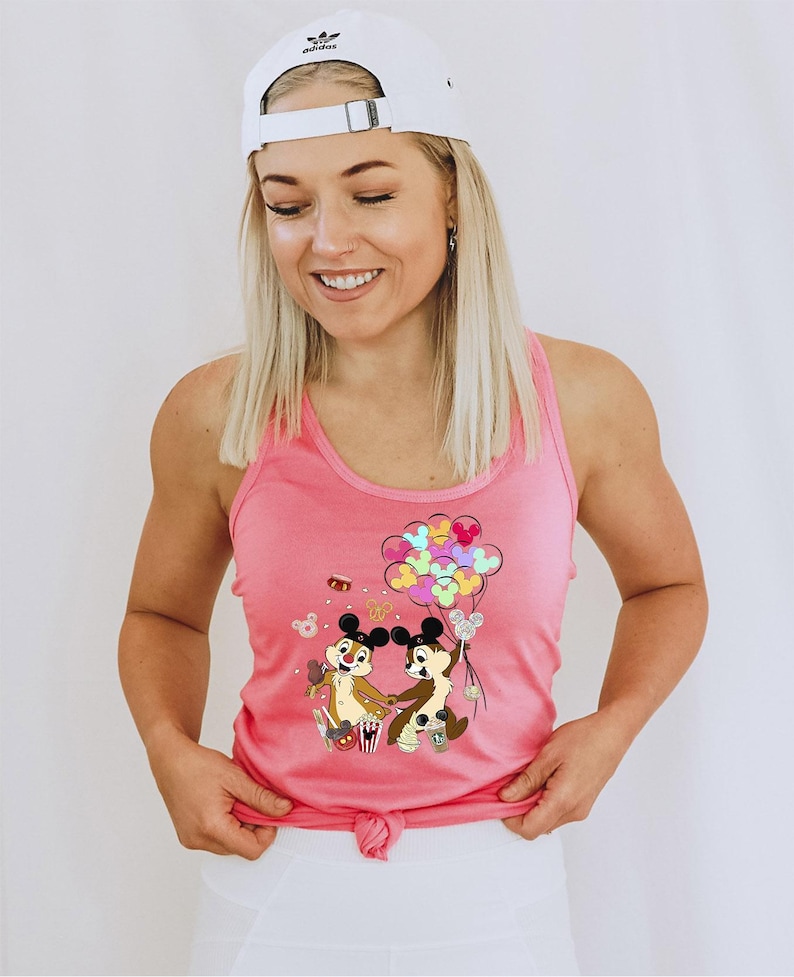 May include: Pink tank top featuring a graphic of Chip and Dale holding hands with balloons. The cartoon characters are surrounded by popcorn and sweets. The top is tied at the waist and worn with a white Adidas baseball cap.