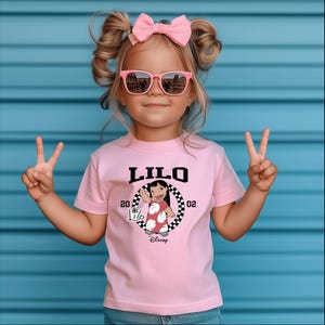 May include: A pink t-shirt featuring the word "LILO" in black block letters, with a graphic of a cartoon character and the text "20 02". The shirt is worn by a young child wearing pink sunglasses and a pink bow.