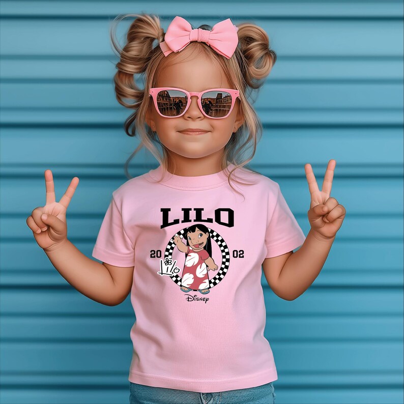 May include: A pink t-shirt featuring the word "LILO" in black block letters, with a graphic of a cartoon character and the text "20 02". The shirt is worn by a young child wearing pink sunglasses and a pink bow.