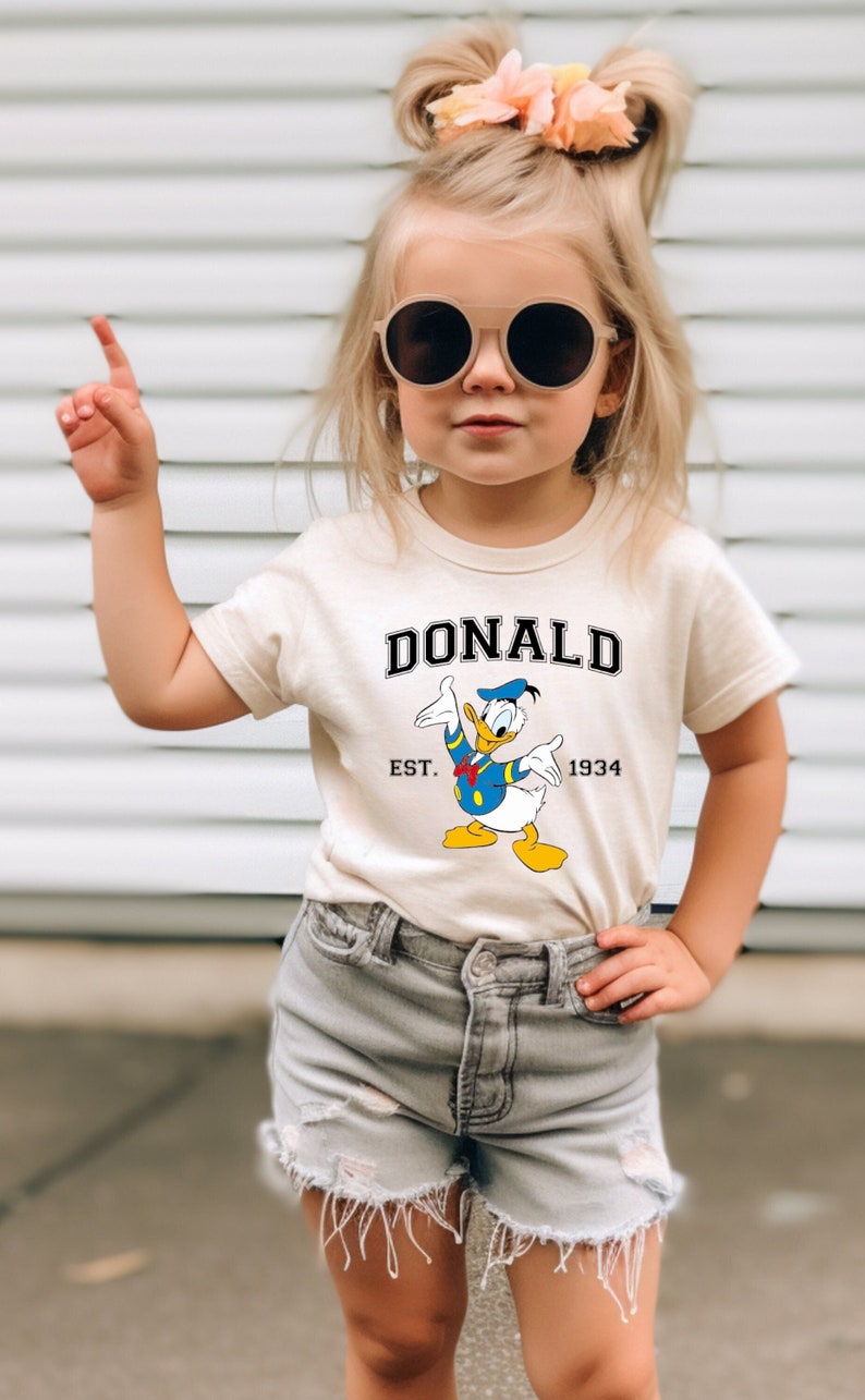 May include: A young girl wearing a white t-shirt with a cartoon image of Donald Duck and the text "DONALD EST. 1934". She is also wearing denim shorts and sunglasses.