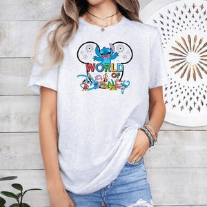 Pu&ograve; includere: Una maglietta grigio chiaro con una grafica colorata di Stitch di Lilo & Stitch, con le parole "WORLD OF DISNEY". Il design include altri personaggi Disney. La maglietta &egrave; indossata da una persona.