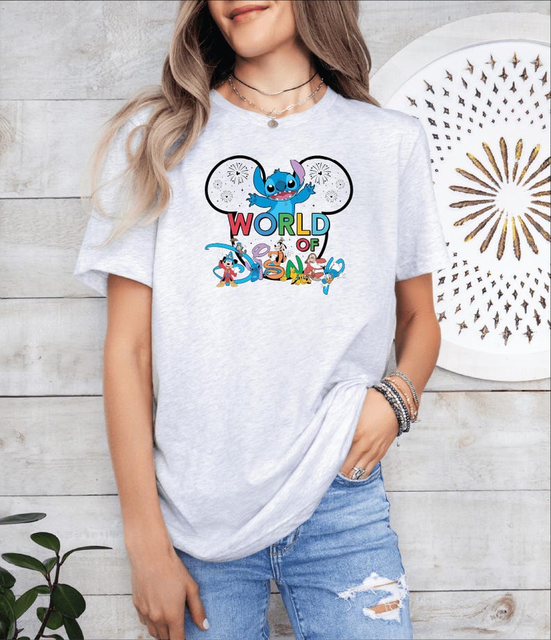 Pu&ograve; includere: Una maglietta grigio chiaro con una grafica colorata di Stitch di Lilo & Stitch, con le parole "WORLD OF DISNEY". Il design include altri personaggi Disney. La maglietta &egrave; indossata da una persona.