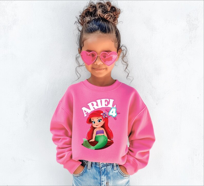 Może przedstawiać: Dziewczynka w r&oacute;żowej bluzie z Arielką z Małej Syrenki. Bluza ma napis "ARIEL" w kolorze białym i cyfrę "4". Dziewczynka ma r&oacute;wnież r&oacute;żowe okulary przeciwsłoneczne w kształcie serca i niebieskie dżinsy.
