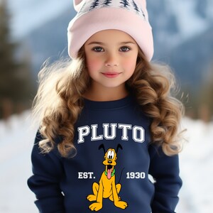 Puede incluir: Una sudadera azul marino con un gr&aacute;fico blanco de Pluto, el perro de Disney. El gr&aacute;fico incluye el texto "PLUTO EST. 1930". La sudadera la lleva puesta una ni&ntilde;a que lleva una gorra de punto rosa con un dise&ntilde;o de &aacute;rbol blanco.
