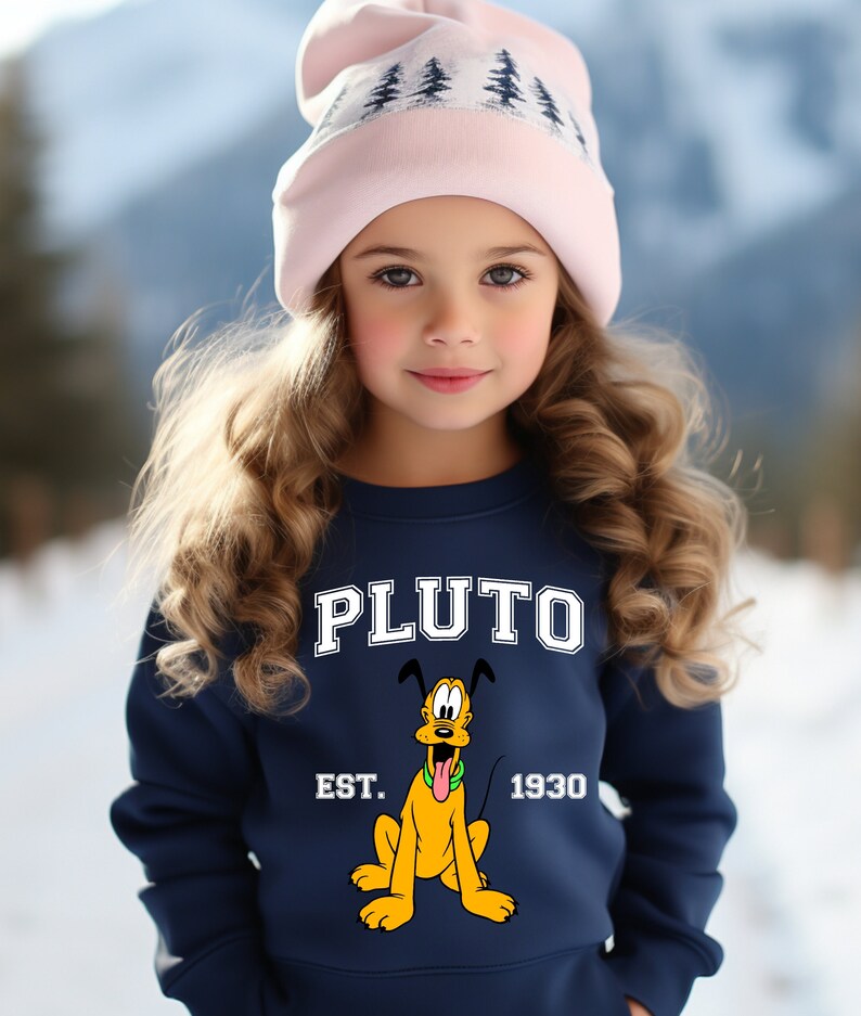 Puede incluir: Una sudadera azul marino con un gr&aacute;fico blanco de Pluto, el perro de Disney. El gr&aacute;fico incluye el texto "PLUTO EST. 1930". La sudadera la lleva puesta una ni&ntilde;a que lleva una gorra de punto rosa con un dise&ntilde;o de &aacute;rbol blanco.