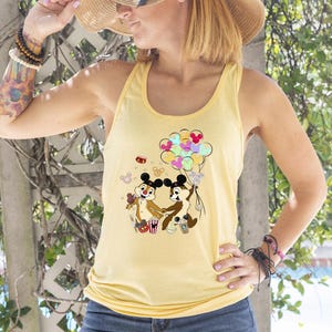 May include: A light yellow tank top featuring a cartoon graphic of Chip and Dale holding balloons. The graphic includes colorful balloons and popcorn. The tank top is sleeveless and has a rounded neckline.