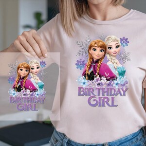 K&ouml;nnte beinhalten: Hellrosa T-Shirt mit einem Aufdruck von zwei animierten M&auml;dchen und dem Text "BIRTHDAY GIRL" in Lila. Die Grafik enth&auml;lt lila und blaue Blumenelemente und Schneeflocken.