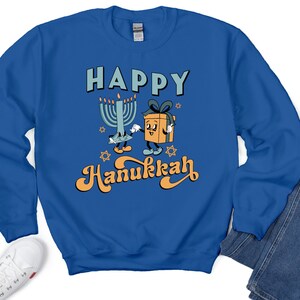 May include: Royal blue sweatshirt with the words "HAPPY Hanukkah" in a retro font. The graphic features a cartoon menorah and a gift box with arms and legs. White sneakers with a leopard print accent are visible.