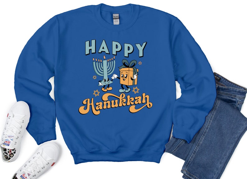 May include: Royal blue sweatshirt with the words "HAPPY Hanukkah" in a retro font. The graphic features a cartoon menorah and a gift box with arms and legs. White sneakers with a leopard print accent are visible.