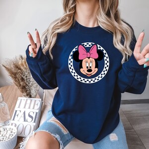 Puede incluir: Una sudadera azul marino con un c&iacute;rculo a cuadros blancos y negros que presenta una imagen de dibujos animados de Minnie Mouse con un lazo rosa.