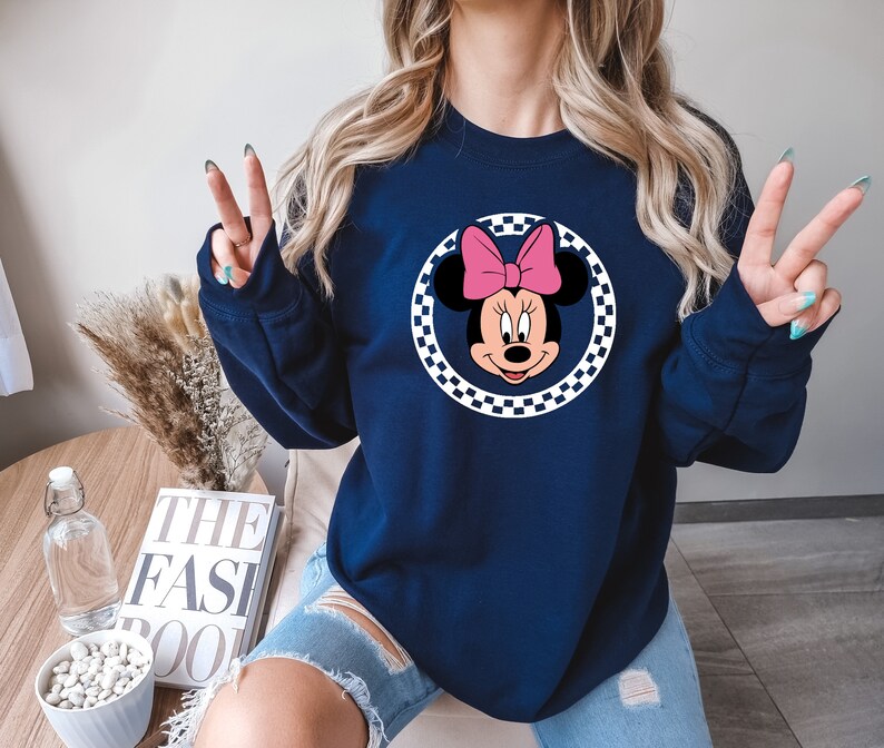Puede incluir: Una sudadera azul marino con un c&iacute;rculo a cuadros blancos y negros que presenta una imagen de dibujos animados de Minnie Mouse con un lazo rosa.