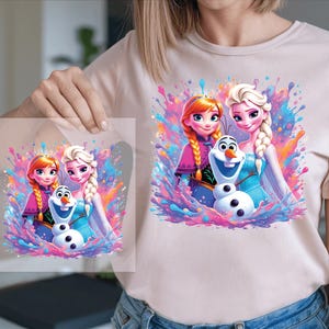 Pu&ograve; includere: Una maglietta rosa chiaro con una grafica colorata di due principesse e un pupazzo di neve. Il design include vivaci spruzzi di rosa, blu e viola. L'immagine &egrave; tenuta da una persona.