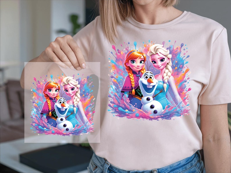 Pu&ograve; includere: Una maglietta rosa chiaro con una grafica colorata di due principesse e un pupazzo di neve. Il design include vivaci spruzzi di rosa, blu e viola. L'immagine &egrave; tenuta da una persona.
