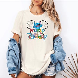 Pu&ograve; includere: T-shirt color crema con grafica colorata di Stitch di Lilo & Stitch, fuochi d'artificio e la scritta "WORLD OF DISNEY" in un font giocoso. Il design &egrave; racchiuso in un contorno di orecchie di Topolino. Parte di una collezione a tema Disney.
