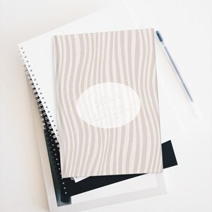May include: A stack of notebooks and a pen on a white surface. The top notebook has a beige and white wavy pattern with the words "DEAR DIARY..." in a white oval. Other notebooks are white and black with spiral bindings.
