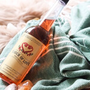 May include: A glass bottle with a pink liquid inside. The bottle has a label that says "Touch of Love Patchouli" with a red heart design. The bottle is sitting on a green and white striped towel.