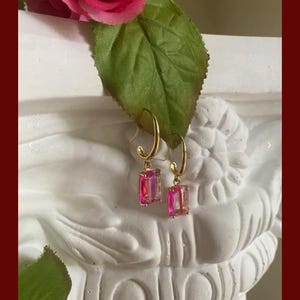 Könnte beinhalten: Goldfarbene Creolen mit rechteckigen, schillernden rosa- und lilafarbenen Kristallen. Die Ohrringe werden auf einer weißen, skulptierten Büste mit einer rosa Rose und einem grünen Blatt präsentiert.