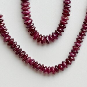 May include: A strand of deep red gemstone beads, likely rubies, arranged in a necklace form. The beads are small, round, and closely spaced, with a matching red tassel at the end. The background is plain white.