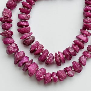 Può includere: Due fili di perline di pietre preziose di rubino grezzo in varie tonalità di rosa e rosso. Le perline sono infilate con un filo bianco e rifinite con nappe rosse. Le pietre preziose hanno una forma irregolare.