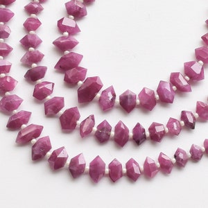 May include: A strand of pink gemstone beads with a faceted, elongated, diamond-shaped design. The beads are strung on a white thread.