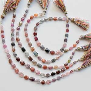 May include: A set of six strands of oval-shaped gemstone beads in various shades of pink, purple, blue, and green. Each strand has a colorful tassel at the end.