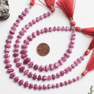 Pu&ograve; includere: Tre fili di perle di rubino rosa con perle sfaccettate a forma di diamante. Le perle sono infilate su un filo trasparente e hanno un fiocco rosso alla fine di ogni filo. Un centesimo di dollaro americano &egrave; incluso per la scala.