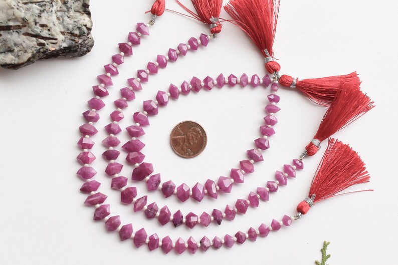 Pu&ograve; includere: Tre fili di perle di rubino rosa con perle sfaccettate a forma di diamante. Le perle sono infilate su un filo trasparente e hanno un fiocco rosso alla fine di ogni filo. Un centesimo di dollaro americano &egrave; incluso per la scala.