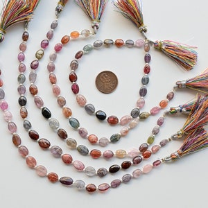 May include: A strand of oval-shaped gemstones in various shades of pink, blue, and green. The gemstones are strung on a silver cord and have colorful tassels at each end.