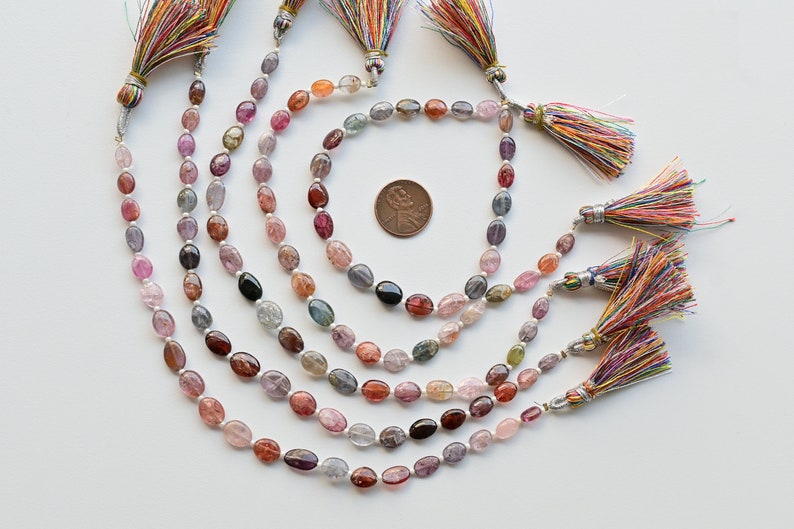 May include: A strand of oval-shaped gemstones in various shades of pink, blue, and green. The gemstones are strung on a silver cord and have colorful tassels at each end.