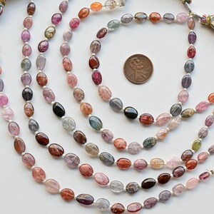 May include: A strand of oval-shaped gemstone beads in various shades of pink, red, blue, and green. The beads are strung on a white cord and are approximately the size of a US penny.
