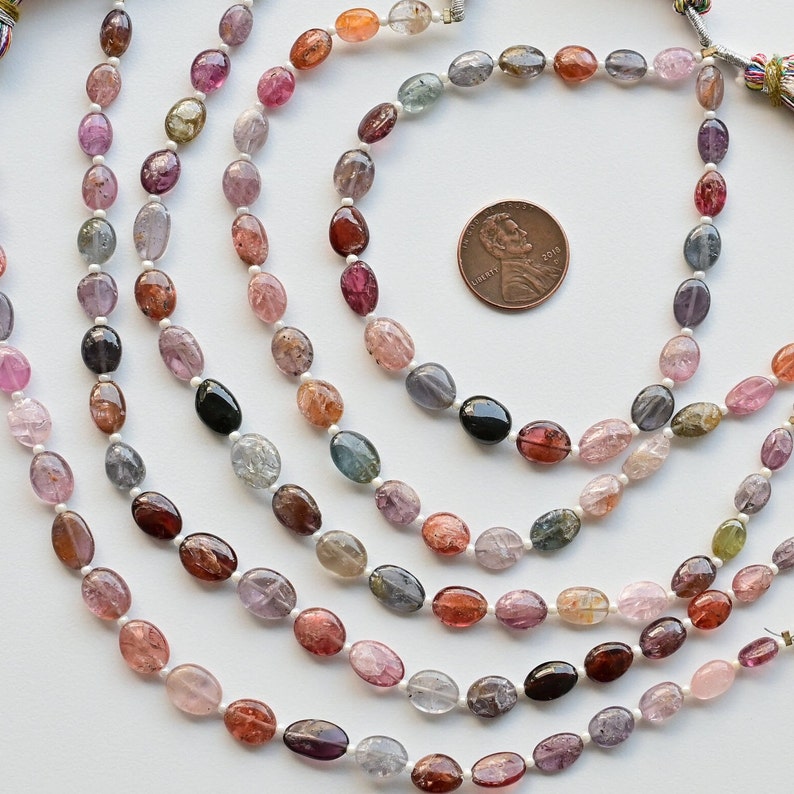 May include: A strand of oval-shaped gemstone beads in various shades of pink, red, blue, and green. The beads are strung on a white cord and are approximately the size of a US penny.
