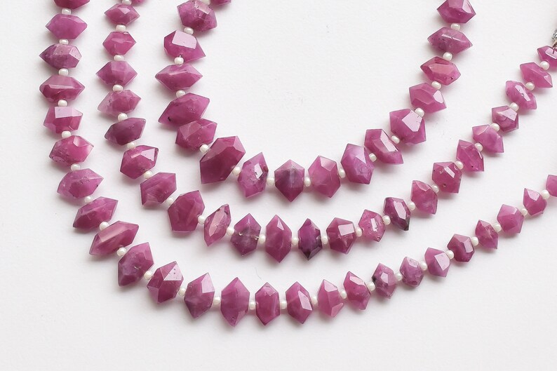 Pu&ograve; includere: Una collana di perline di pietre preziose rosa con un design sfaccettato, allungato, a forma di diamante. Le perline sono infilate su un filo bianco.