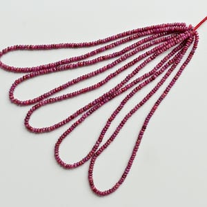 Puede incluir: Hebras de pequeñas cuentas de rubí facetadas. Las cuentas son de un color rojo rosado intenso y rico y están ensartadas en un hilo rojo. Las hebras están dispuestas en forma de abanico sobre un fondo blanco. Adecuado para la fabricación de joyas.