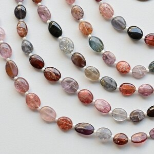 May include: A strand of oval-shaped gemstones in various colors, including pink, red, blue, green, and black. The gemstones are strung on a white thread.