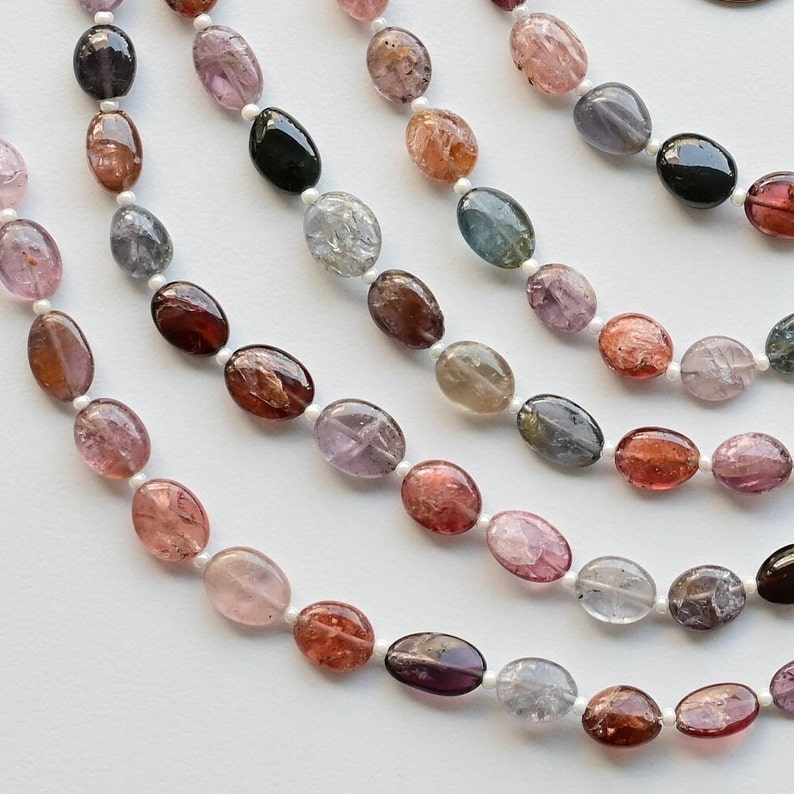 May include: A strand of oval-shaped gemstones in various colors, including pink, red, blue, green, and black. The gemstones are strung on a white thread.