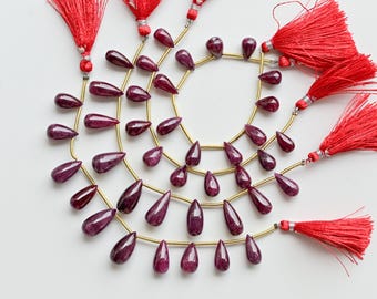 May include: A collection of deep red, teardrop-shaped gemstones strung on gold-coloured wire, arranged in a circular pattern. Bright red tassels with small beads are attached. The gemstones have a polished, reflective surface.