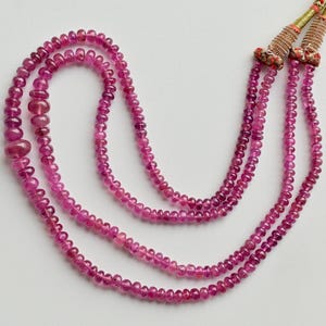 May include: A three-strand necklace featuring small, round, deep pink beads. The necklace has a decorative clasp with gold-colored accents and tassels. The beads are strung together, creating a layered effect. The necklace is displayed on a white background.