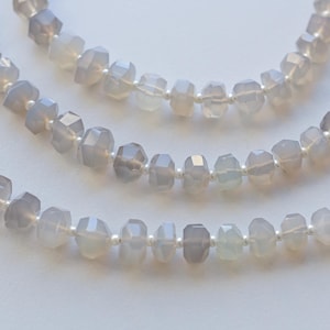 May include: Three strands of faceted, light grey gemstone beads. The beads are strung on a white cord, creating a layered necklace effect. The gemstones have a subtle, translucent quality, reflecting light.