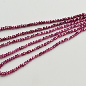 May include: Several strands of small, faceted ruby beads in varying lengths. The beads are a deep, rich pinkish-red colour. The strands are arranged on a white surface, with a red string holding them together at one end.